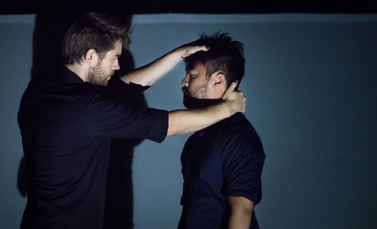Two male performers hold one another in a shadowed blue stage scene.